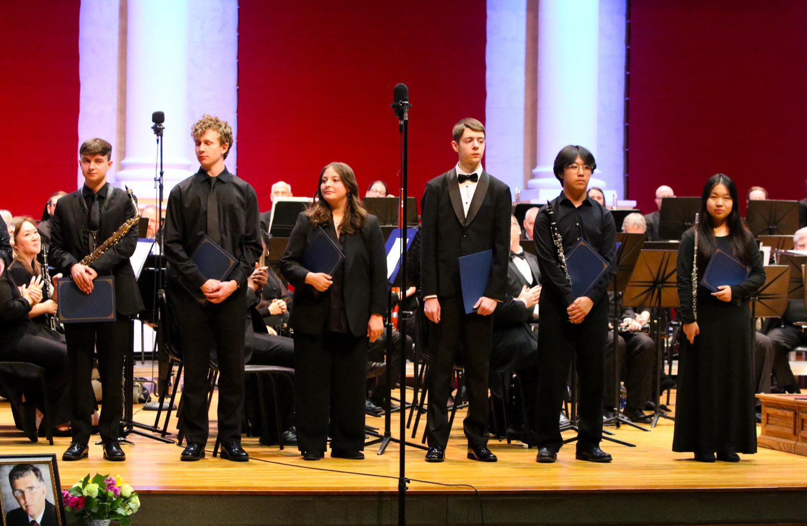 Six students in concert black standing across the stage with their award folders, the band seated behind them